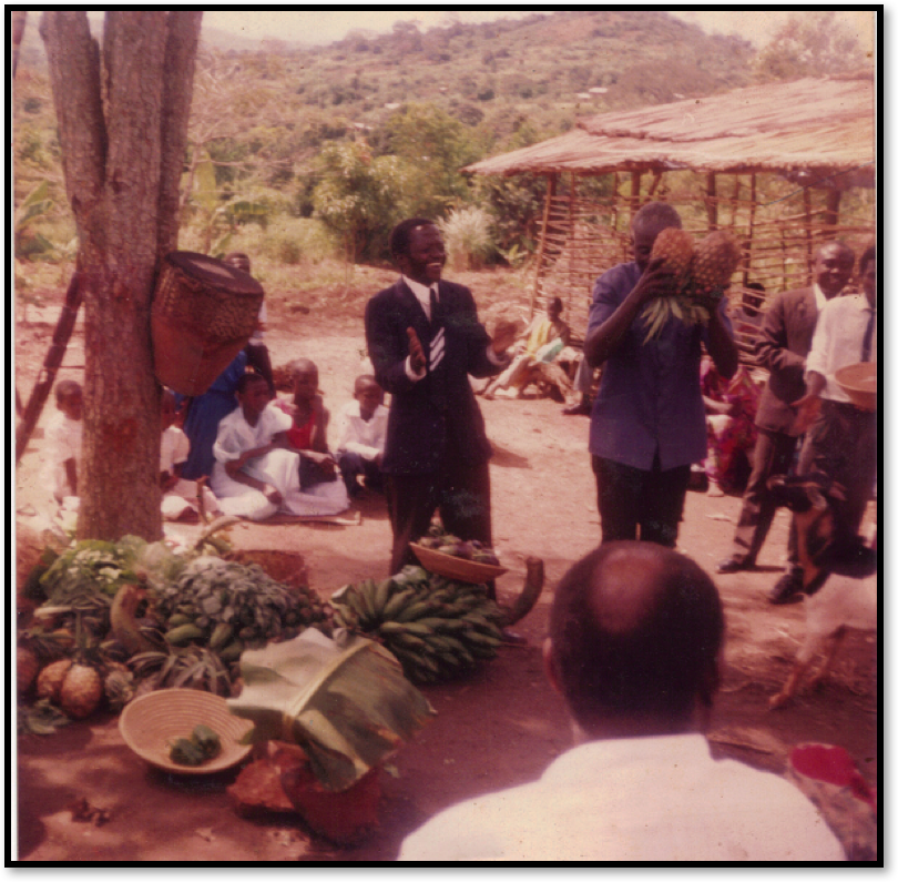 Fundraising after the service outside the church. In this picture: sitting with the back to the camera was Ddamba Benjamin (RIP), holding pineapples was Mr.Kyobe Livingstone, clapping hands was Lay Reader Sebina Lezeni (RIP). On the left hand of the picture is the historical “Mugavu “ tree under which the first service was held on 10.10.1993.